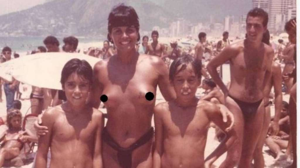 Foto antiga mostra Monique Evans de seios de fora em praia lotada e ao lado de duas crianças